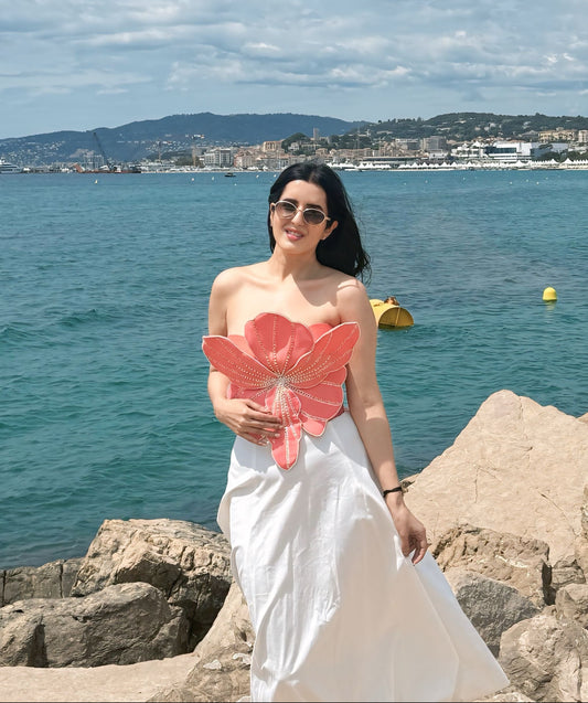 A woman wearing a white strapless maxi dress and sunglasses stands on rocks by the sea, holding a large pink flower-shaped prop. The background features a scenic coastal view with blue water, distant hills, and a cloudy sky.