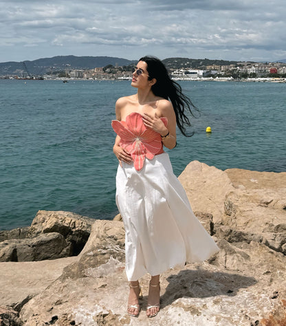 A woman stands on rocky shoreline with the sea and cloudy sky behind her, wearing a strapless white dress and holding a large pink flower.