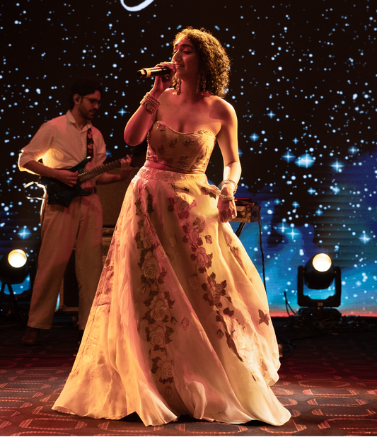 A woman in a strapless cream gown with floral embroidery stands on a stage holding a microphone, with a male musician in the background. Large white text at the top reads 'Sanjeeta BHATTACHARYA' against a starry blue backdrop.