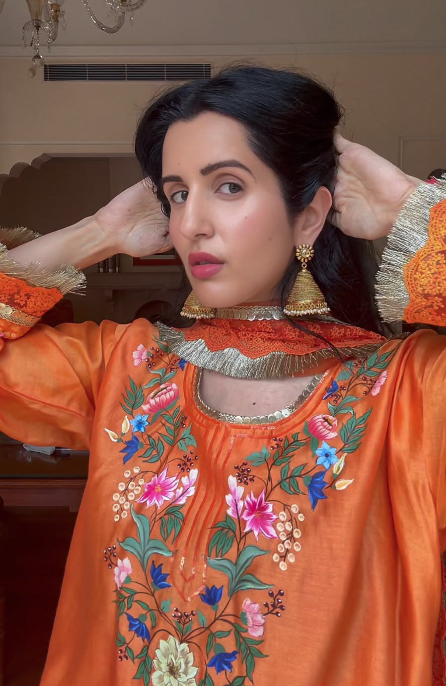 A woman wearing a vibrant orange embroidered traditional outfit with floral patterns, gold earrings, and her hair styled up, posing indoors with a chandelier visible in the background.