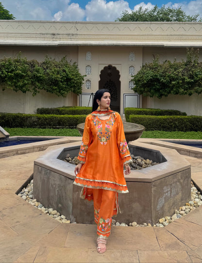 A woman wearing a bright orange embroidered traditional outfit stands in front of a stone fountain in an outdoor courtyard with a beige building, greenery, and a blue sky with clouds in the background.