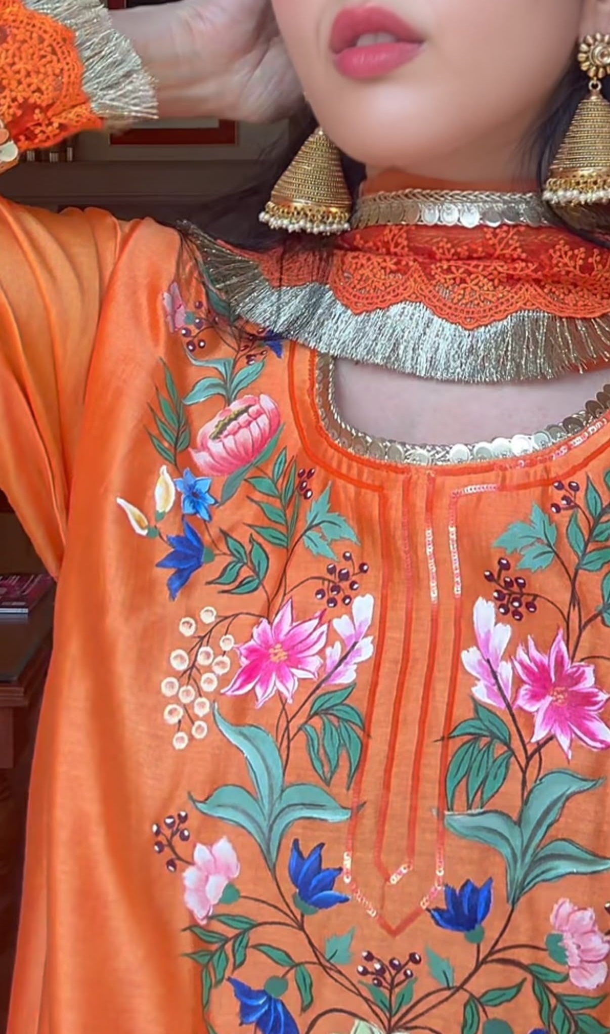 A close-up of a person wearing a bright orange embroidered traditional garment with floral patterns in pink, blue, green, and white, paired with a metallic gold choker and large gold earrings.