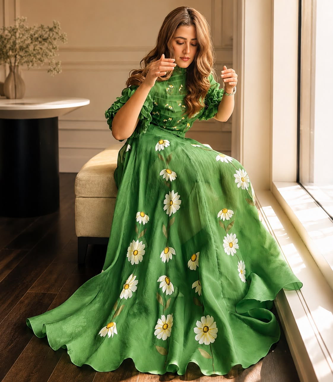 A woman wearing a flowing green dress with white daisy floral prints sits on a beige bench in a sunlit room with dark wood floors and a window.